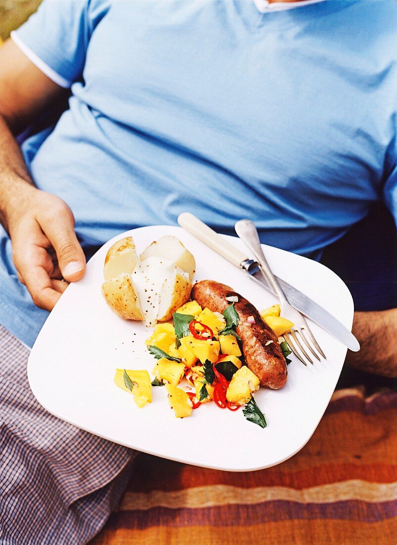 Mann hält Teller mit Grillkartoffel, Salat & Bratwurst