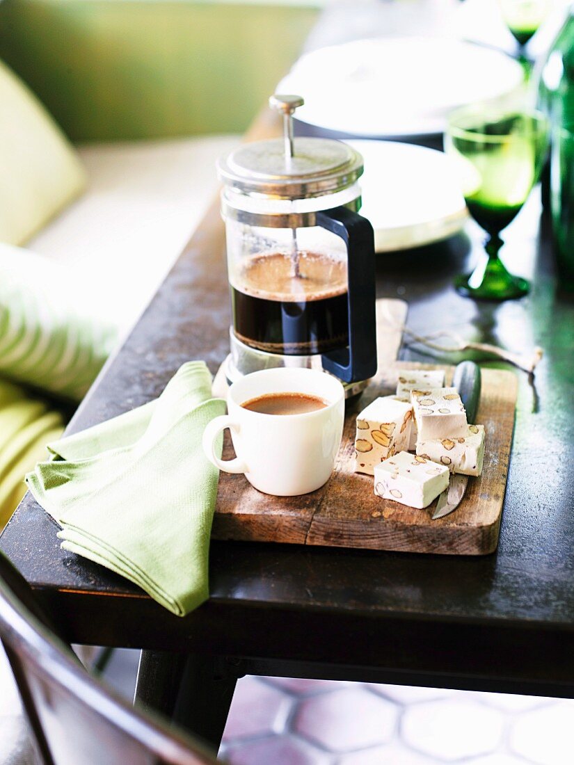 A cafetiere, coffee cups and nougat cubes on a wooden board on a table