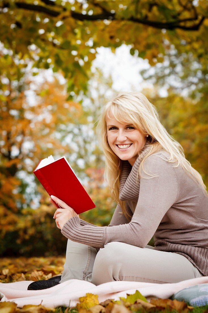 Junge Frau liest ein Buch im Freien auf einer Decke neben Herbstlaub