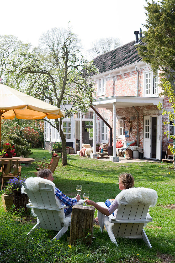 Ehepaar auf Holzsesseln mit Schaffell beim Weintrinken im Garten eines historischen, holländischen Landhauses