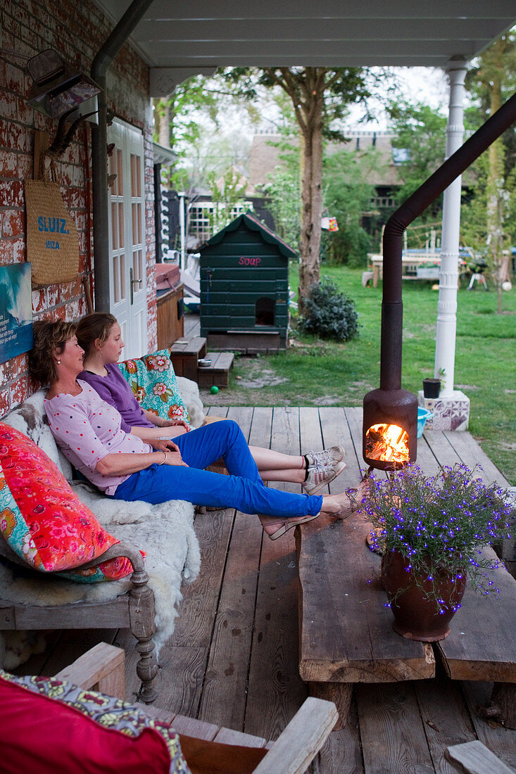 A mother and daughter sitting on a rustic wooden bench in front of a fire on a terrace