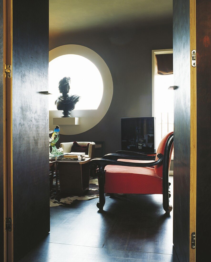 View through door into interior in dark shades with red armchair and sculpture in round window