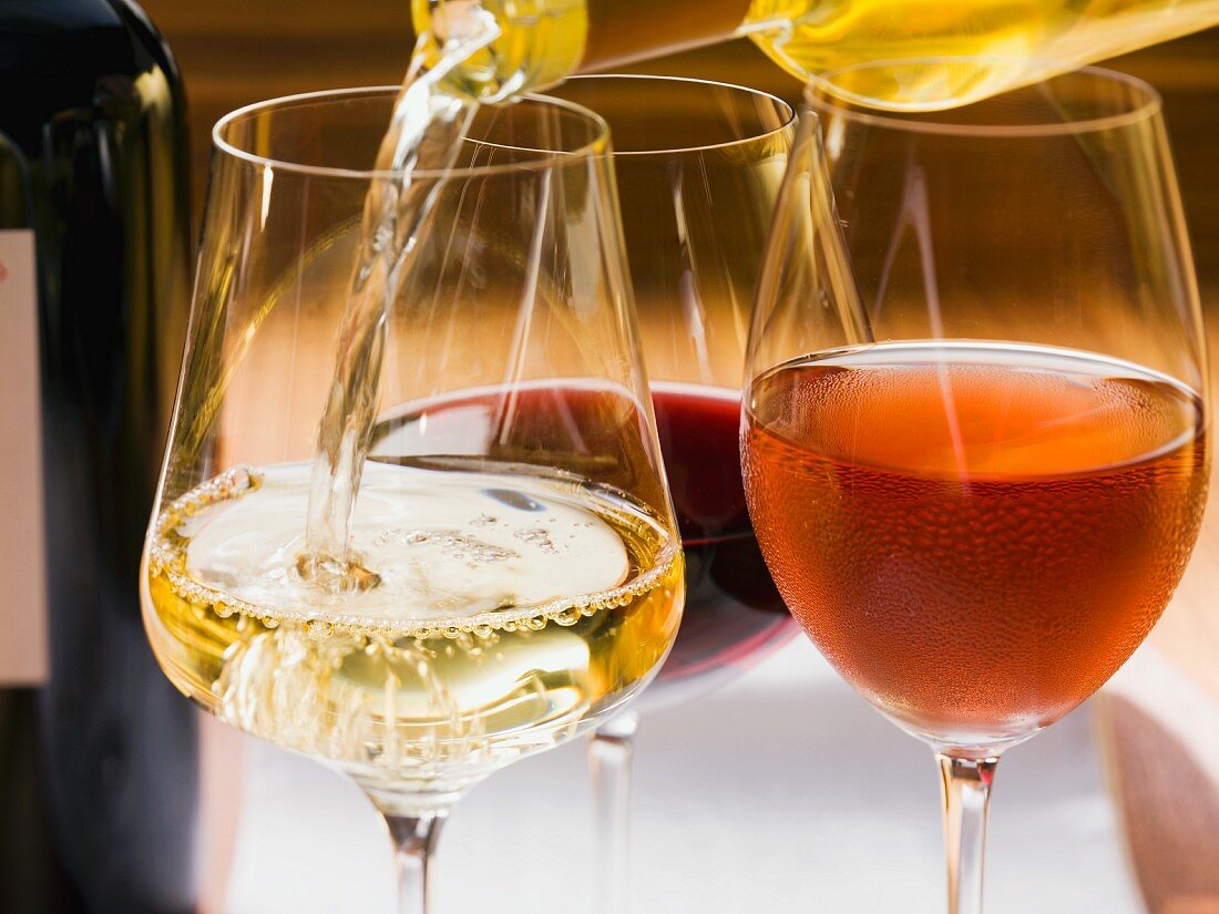 White wine being poured into a glass next to a glass of rose and red wine