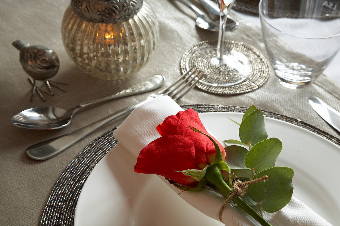Festive place setting with silver accessories and red rose on rolled napkin