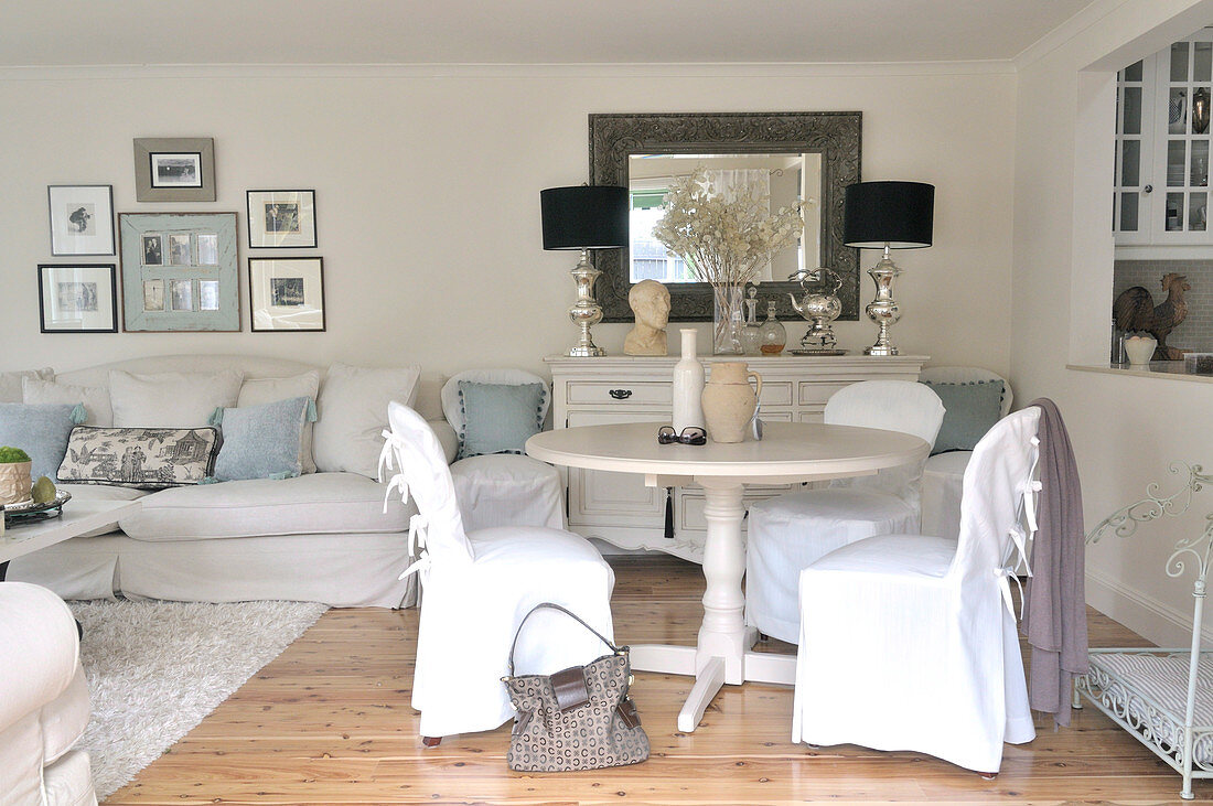 White living room in romantic, elegant country house style with loose-covered chairs at round dining table next to serving hatch leading to kitchen