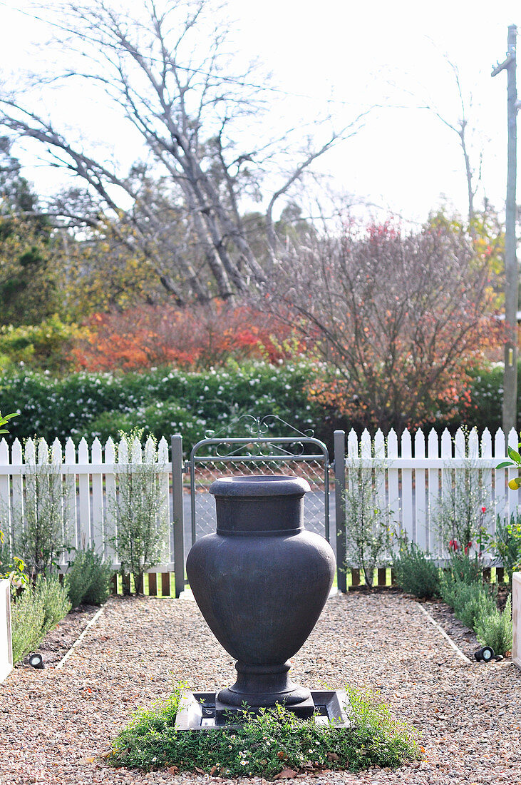 Fountain made from anthracite-coloured amphora on gravel garden path and view of road beyond white wooden fence