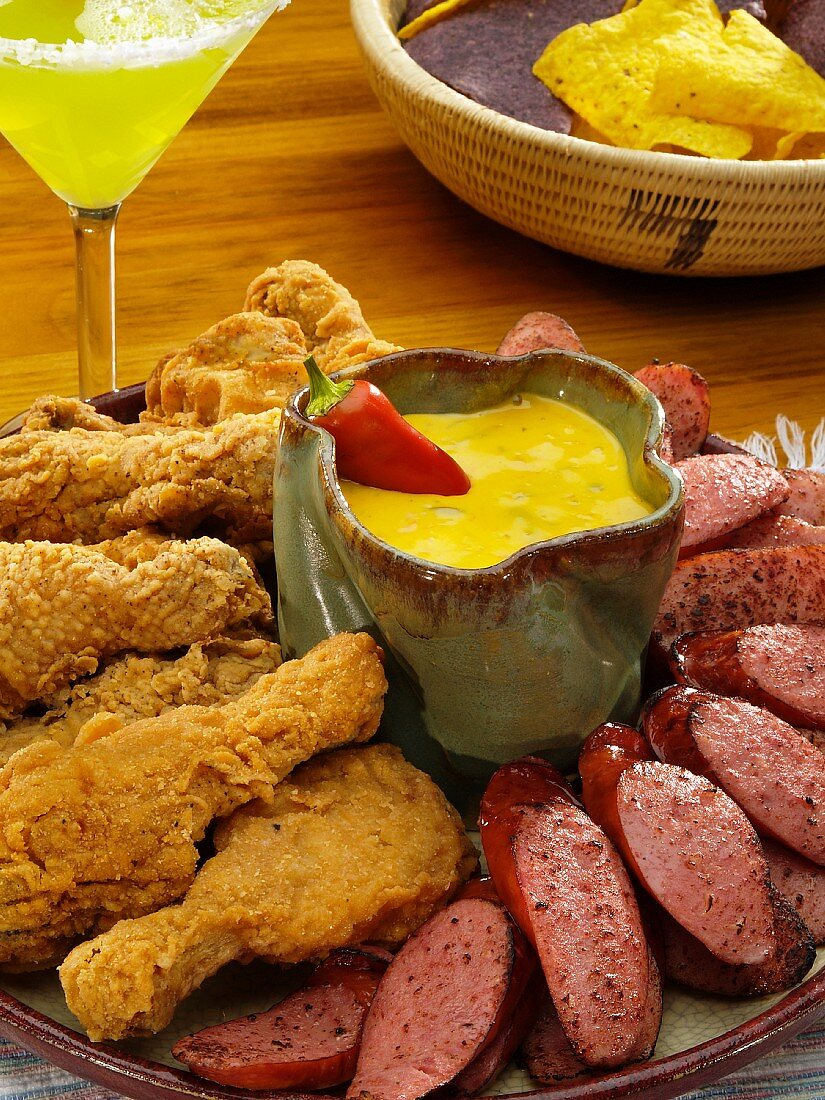 Frittierte Hähnchenschlegel und Wurstscheiben (Kielbasa) mit Tex-Mex-Dip, im Hintergrund Nachos und Margarita