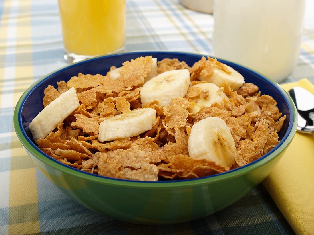 Eine Schale Cornflakes mit Bananen, im Hintergrund Milchflasche und Orangensaft