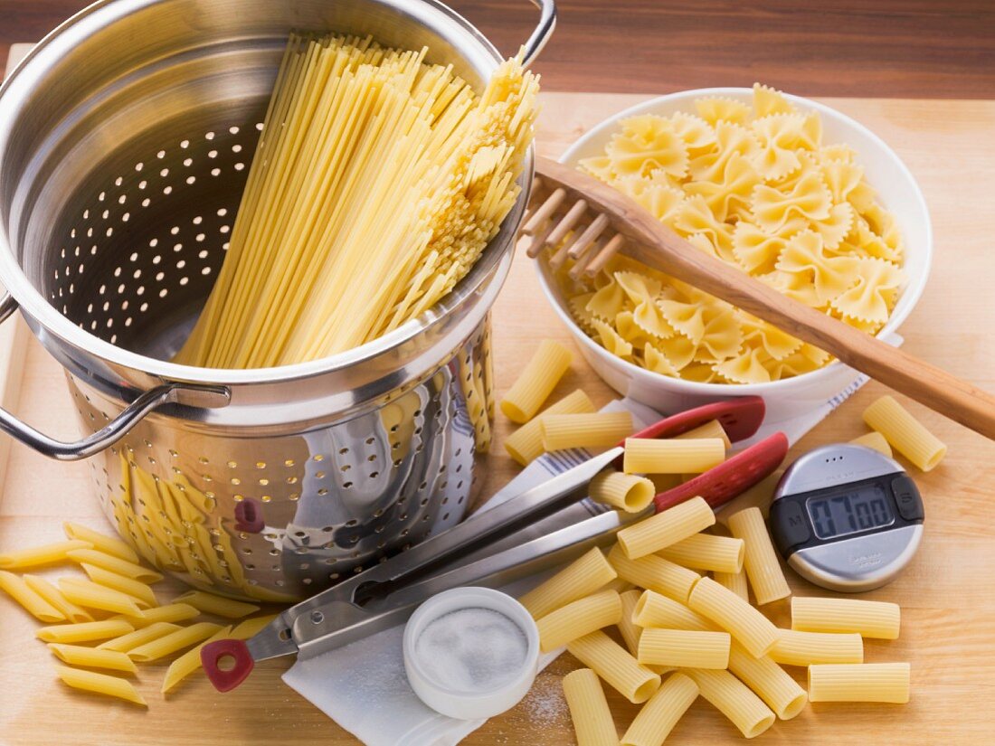 Various types of pasta, a colander, a spaghetti server, tongs and a kitchen timer