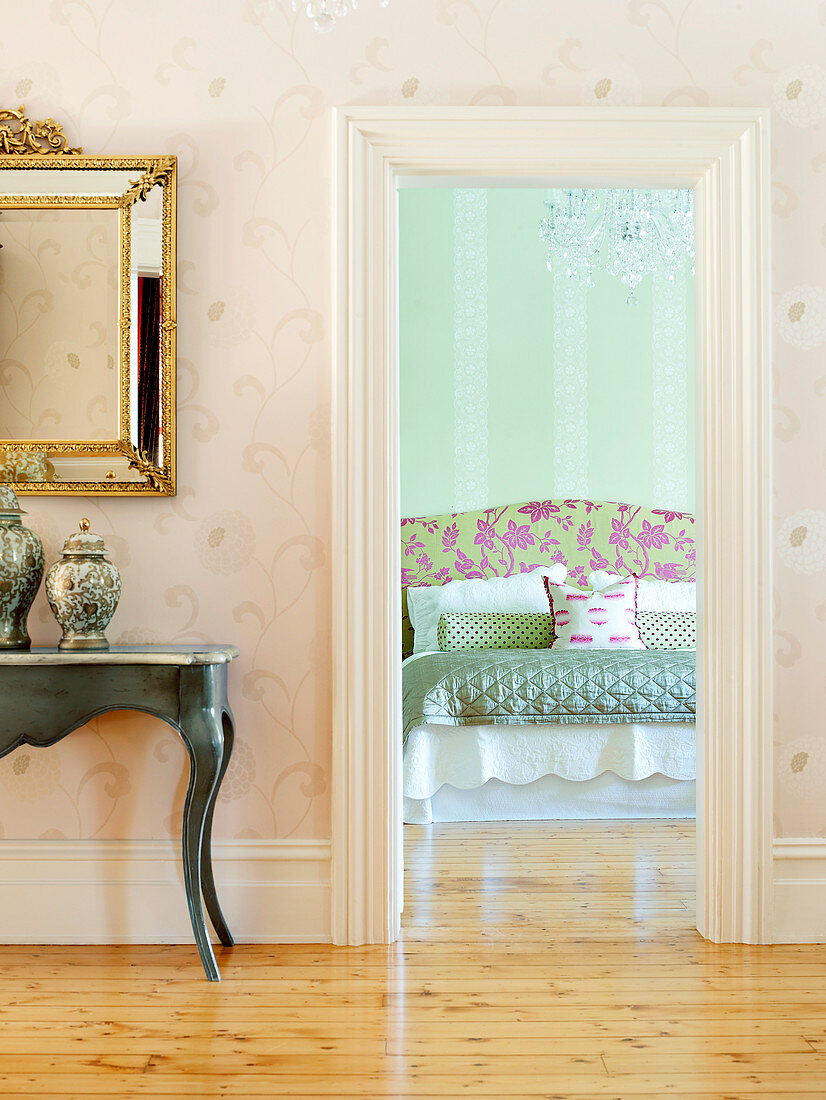 View into bedroom with extravagant double bed below crystal chandelier; gilt-framed mirror above console table in hall