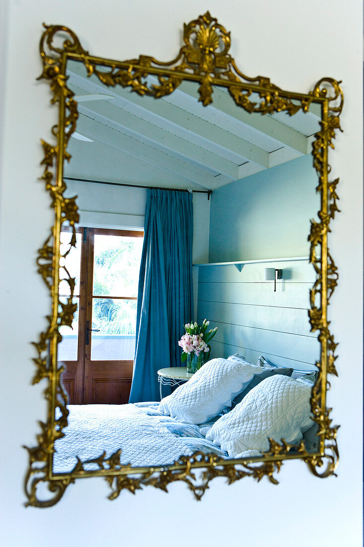 Wall-mounted mirror with floral, gilt ornamented frame reflecting bed in front of French windows with blue curtains