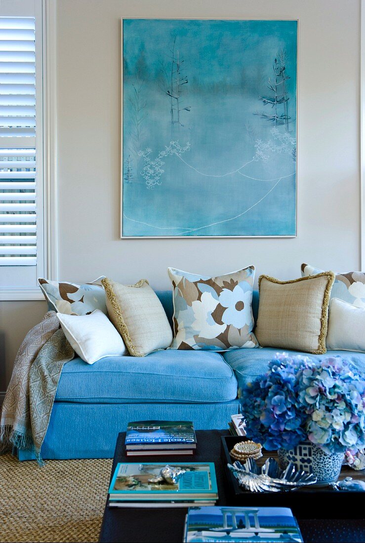 Various scatter cushions on sofa upholstered in light blue below picture on wall and behind flowers on coffee table