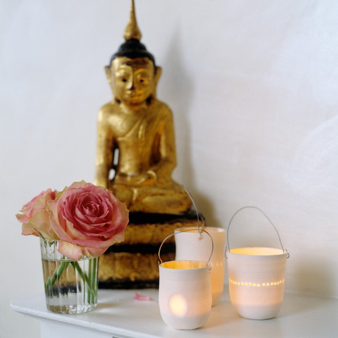 Lit candle lanterns next to roses in glass and gilt Buddha figurine on shelf