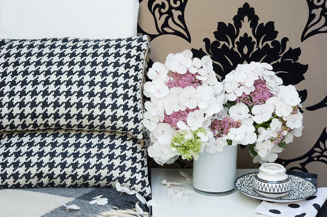 White hydrangeas in small vase next to scatter cushions and against brocade wallpaper