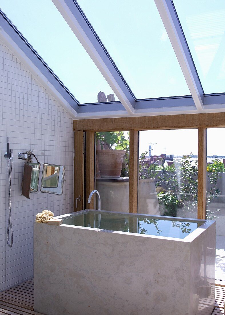 Kubische Badewanne mit Wasser gefüllt unter verglaster Dachschräge und Blick durch Terrassenfenster