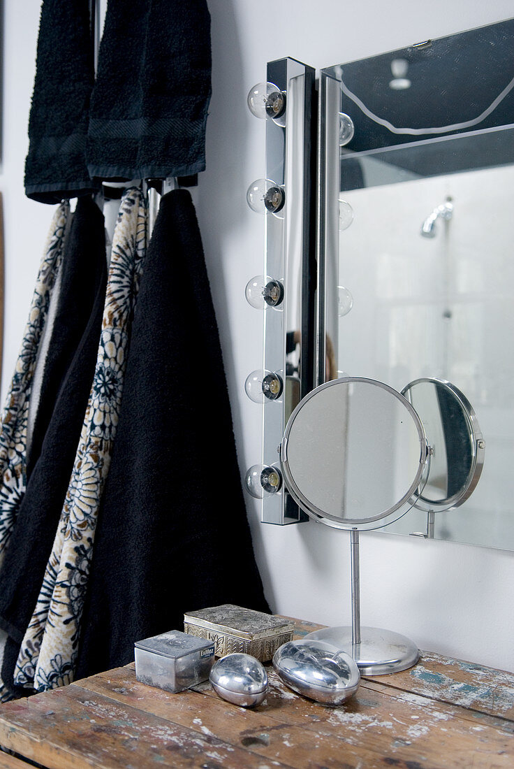 Moderner Badezimmerspiegel mit seitlicher Lichtleiste, alter Tisch mit Silberdöschen und schwarze Handtücher an der Wand hängend