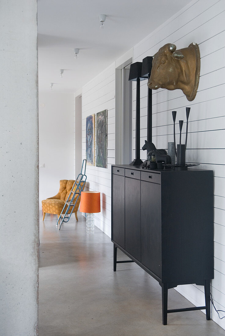 Goldfarbene Kuhkopfskulptur an der Wand über Sideboard mit Deko in einheitlichem Schwarz; Retro-Sessel und Tretleiter im Hintergrund