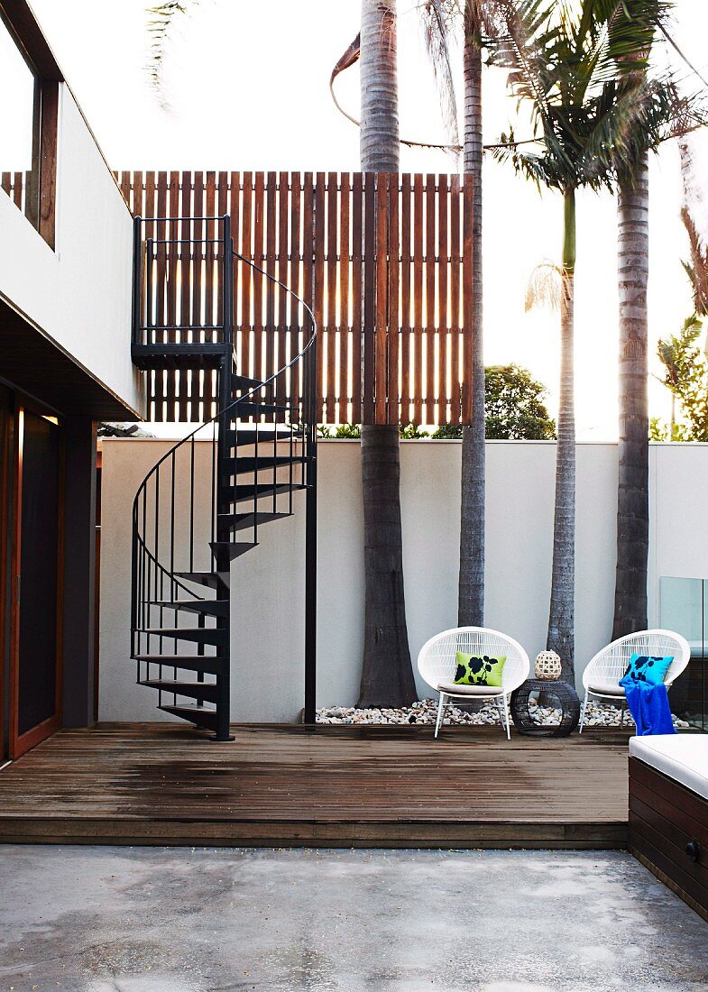 Stylish courtyard of contemporary house with white wicker chairs on wooden platform in front of palm trees and spiral staircase