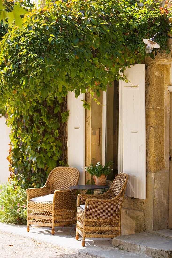 Wicker chairs and side table in front of windows with white wooden shutters in climber-covered house facade