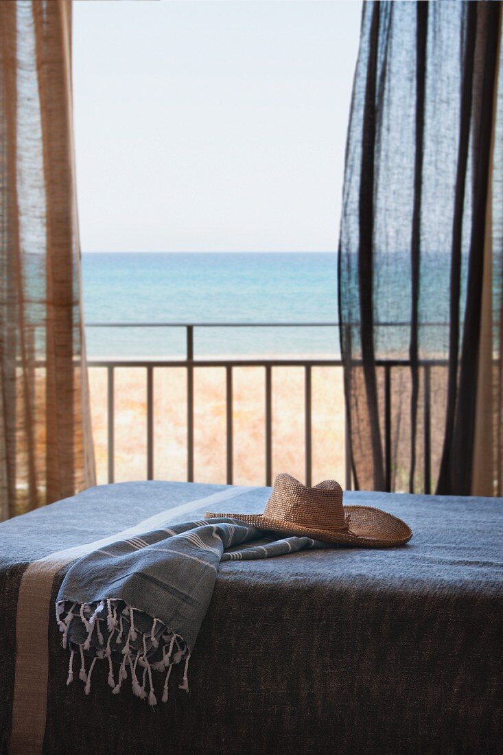 Straw hat on day-bed in front of open balcony door with fluttering curtains and sea view