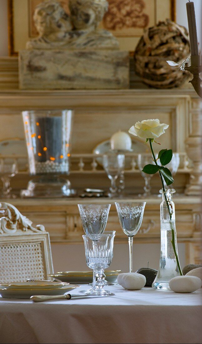 Table set in pale colours with etched glasses, pebbles and white rose; natural wood antique dresser in background