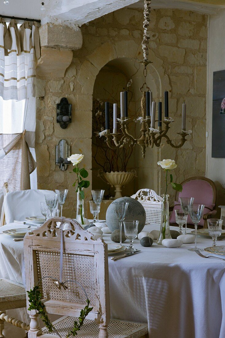 Brass chandelier above festively set table with antique, white, vintage-look chairs; stone wall in background
