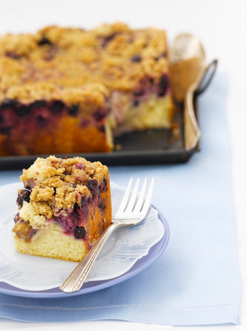 Piece of Blueberry Buckle Cake on a Plate with a Fork; Whole Cake in Background