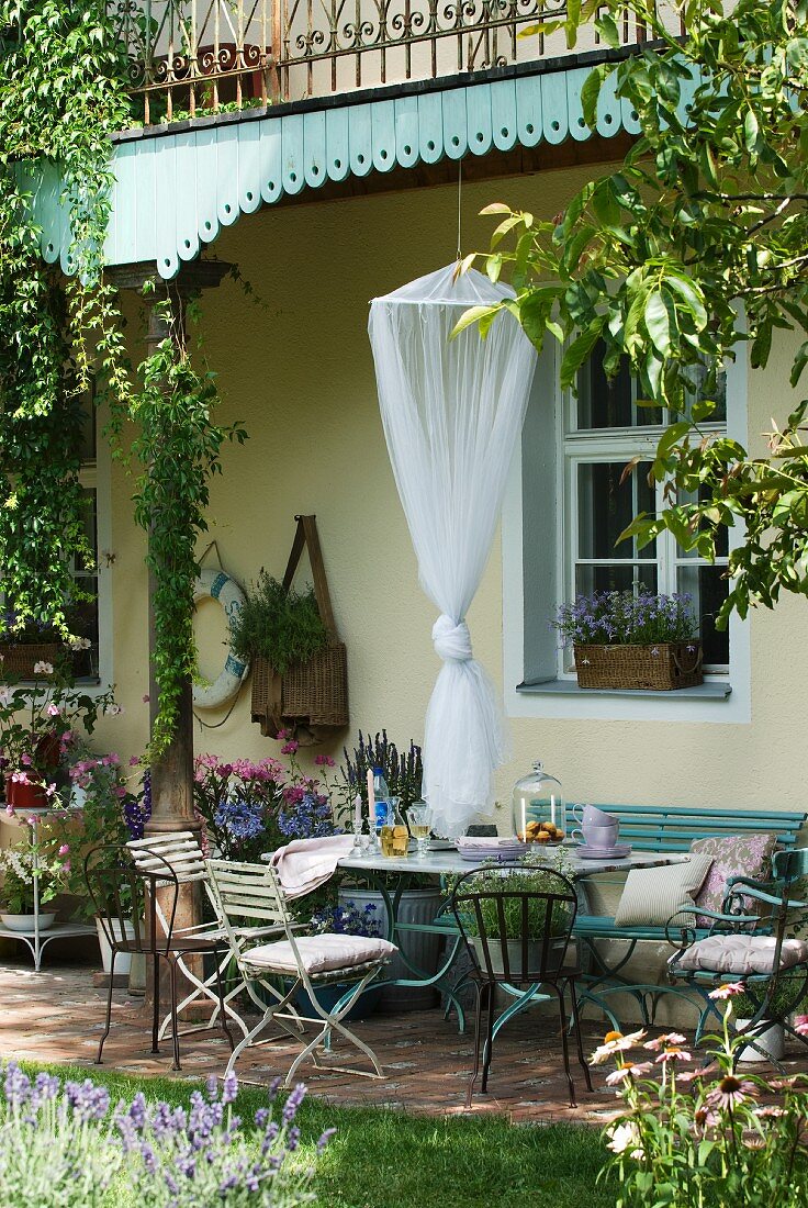 Verschiedene Gartenmöbel im Bistro-Stil und verknotetes Moskitonetz unter verschnörkeltem Balkongeländer und pastellblauer Holzverschalung