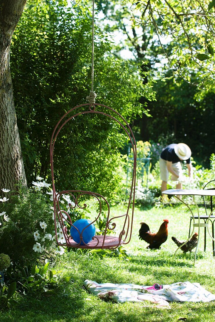 Gartenidylle - Drahtstuhl an einem Seil im Baum hängend, Hühner unter Bistrotisch und arbeitende Person im Hintergrund