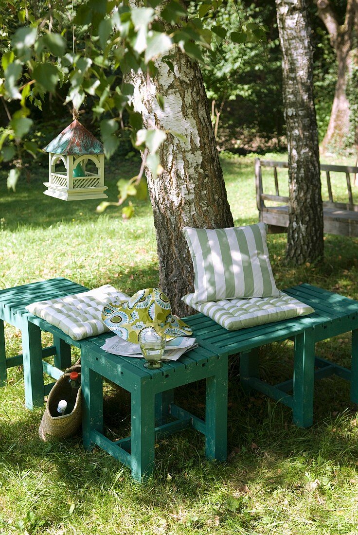 Seating area with striped cushions on small, green wooden bench against birch tree