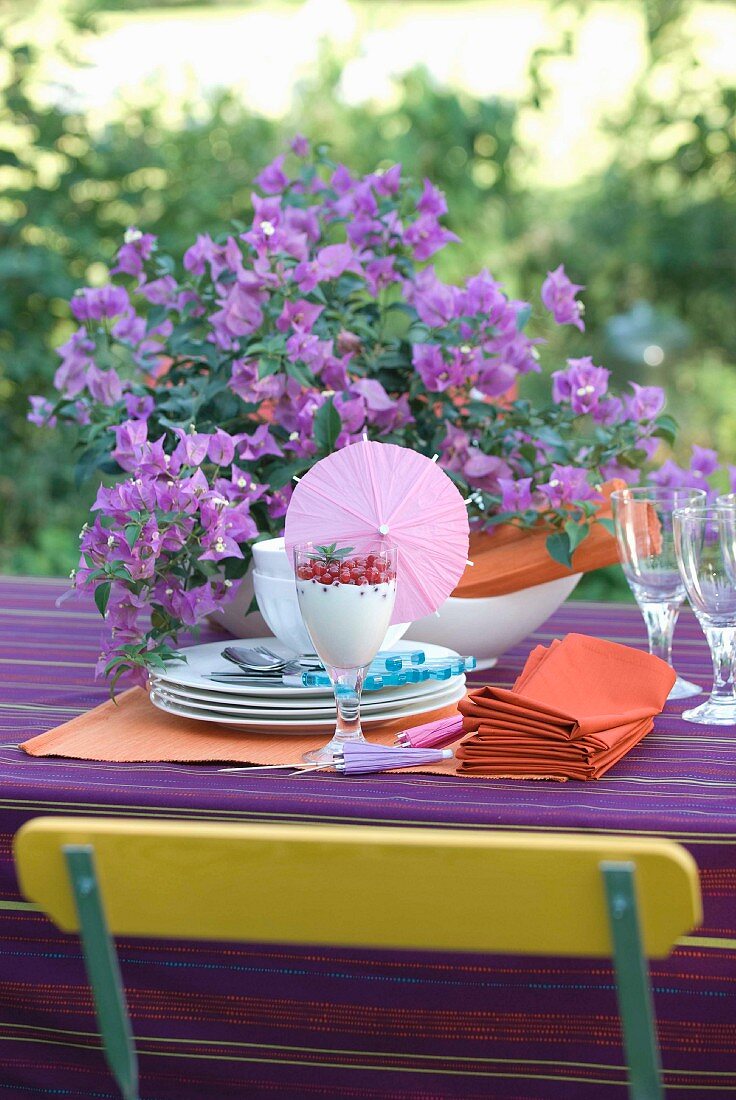 Vorbereitungsstadium eines sommerlich gedeckten Gartentisches mit Bougainvillea auf violettem Tischtuch und Stuhllehne in der Komplementärfarbe Gelb