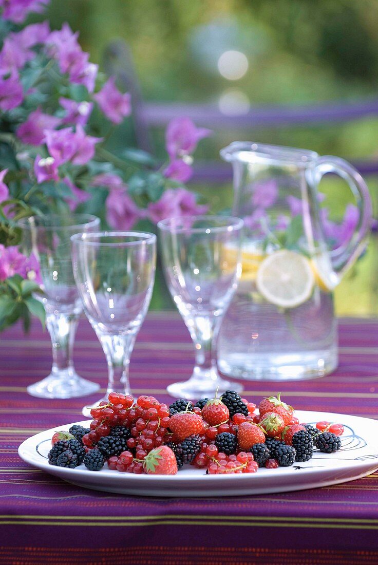 Mixed summer berries on striped purple tablecloth in front of water jug and glasses with bougainvillea flowers in hazy background