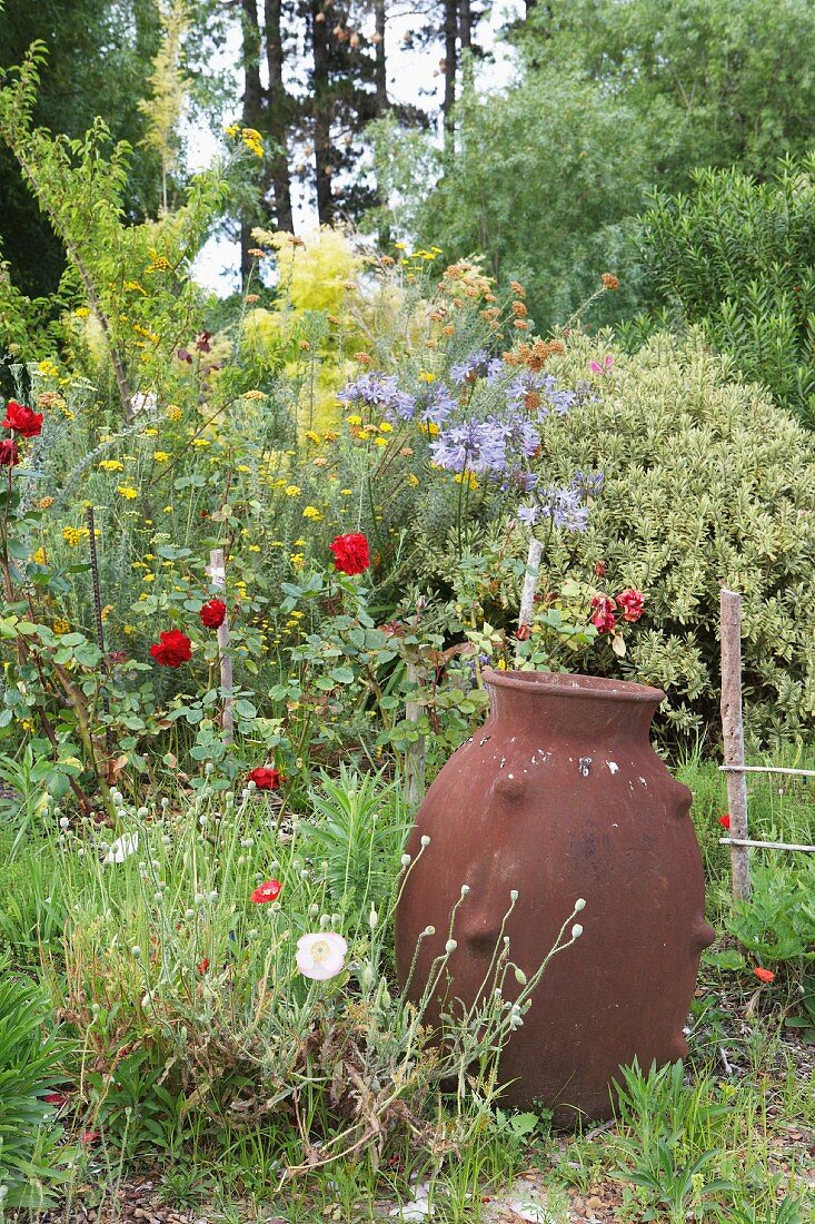 Clay amphora in front of flower bed with a riot of growth and trees in the background