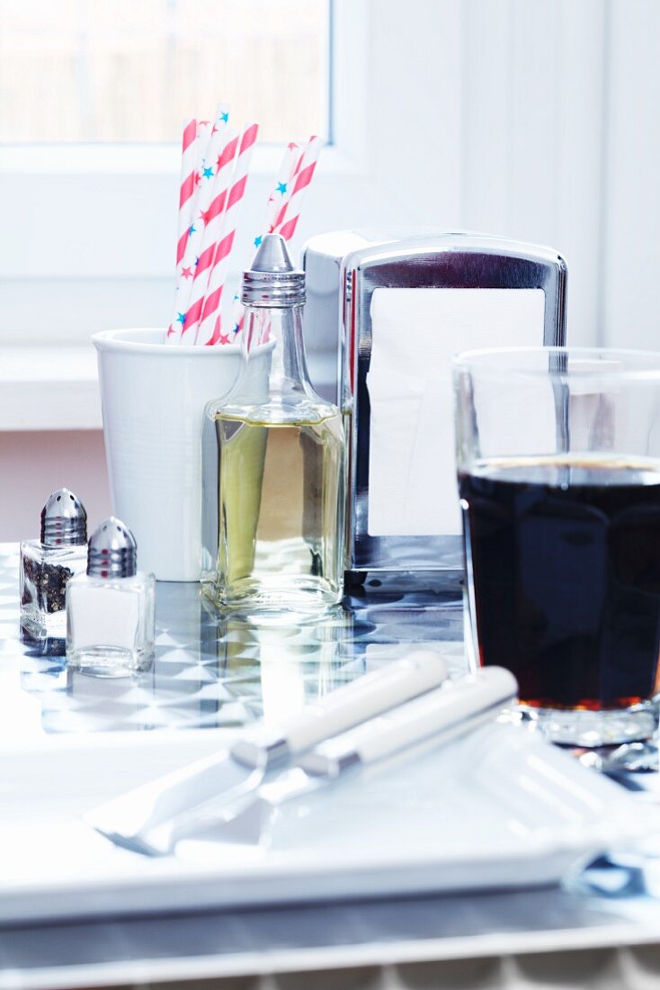 A bottle of vinegar, a napkin dispenser, straws, salt and pepper shakers, a place setting and a glass of cola on a table