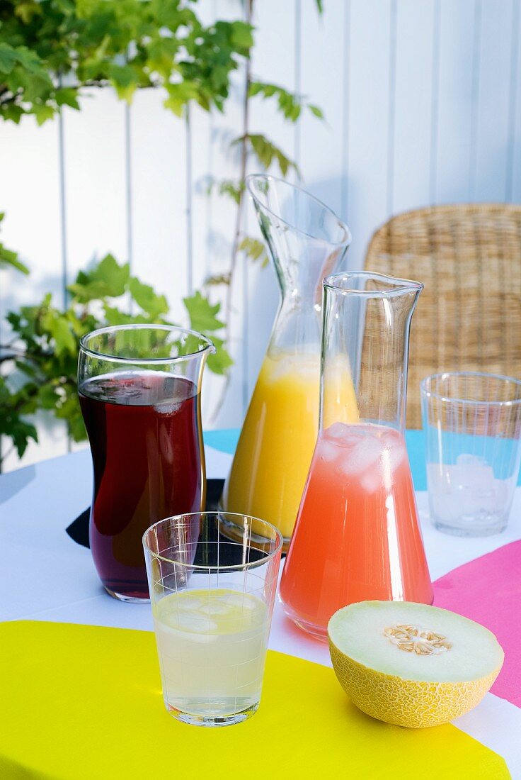 Mit Eiswürfeln gekühlte Fruchtsaftdrinks in Glaskaraffen auf bunt gedecktem Sommertisch im Garten