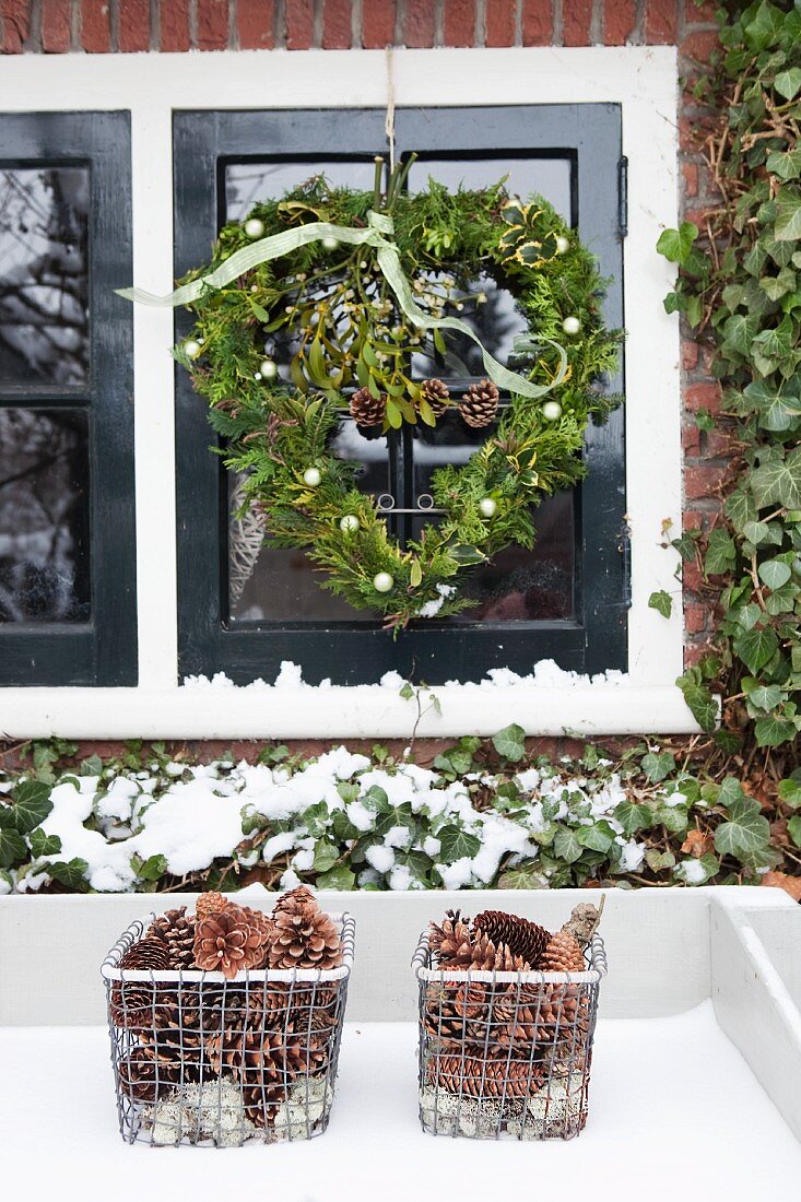 Detail of facade - heart-shaped wreath hanging in front of window and storage baskets of pine cones in snow