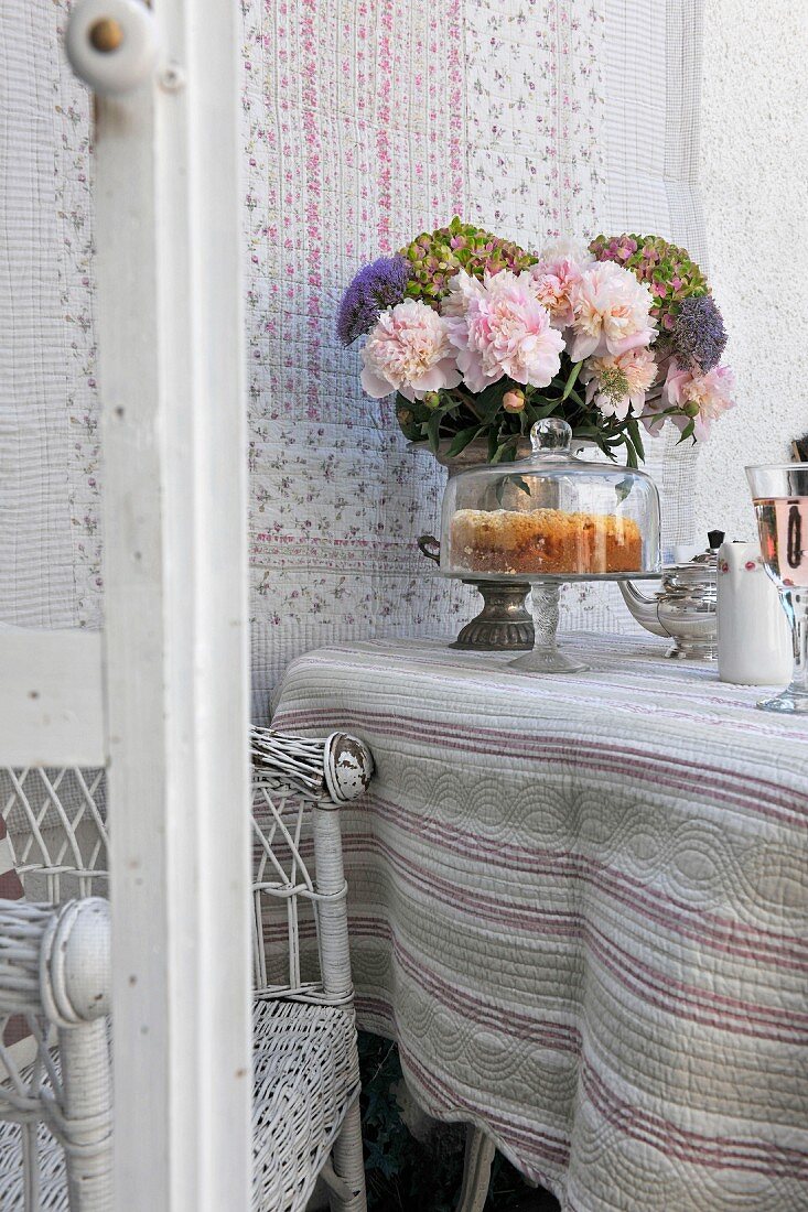 Folkloristisches Tischplaid auf Tisch mit Kuchen unter Glashaube und Blumenstrauss vor Wandbehang