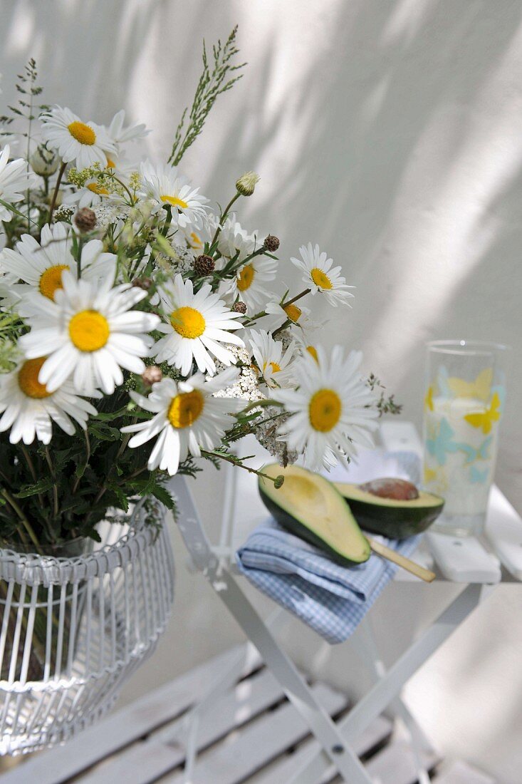 Bouquet of ox-eye daisies next to cut avocado on garden chair