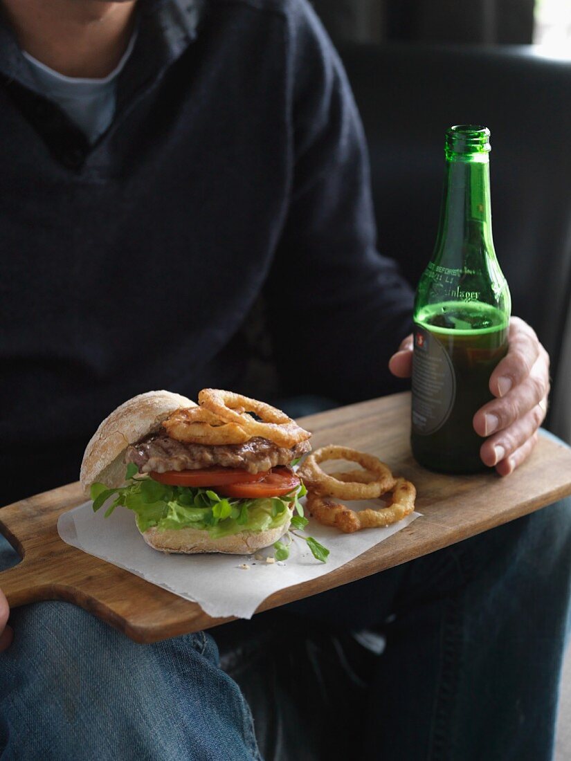 A steak burger with onion rings and a bottle of beer on a wooden board
