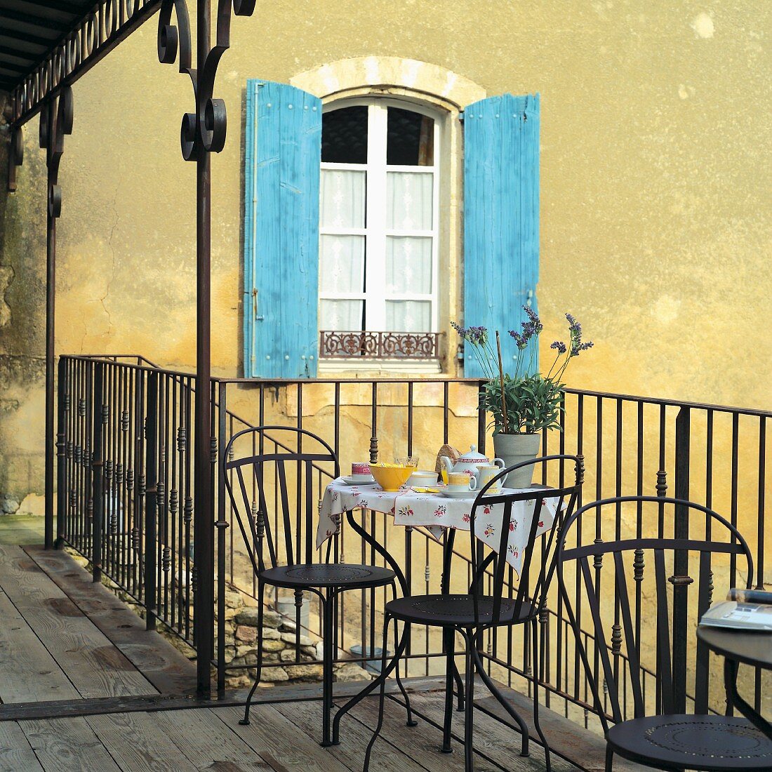 Metal bistro table set for two on wooden balcony with simple metal railing; blue shutters in background