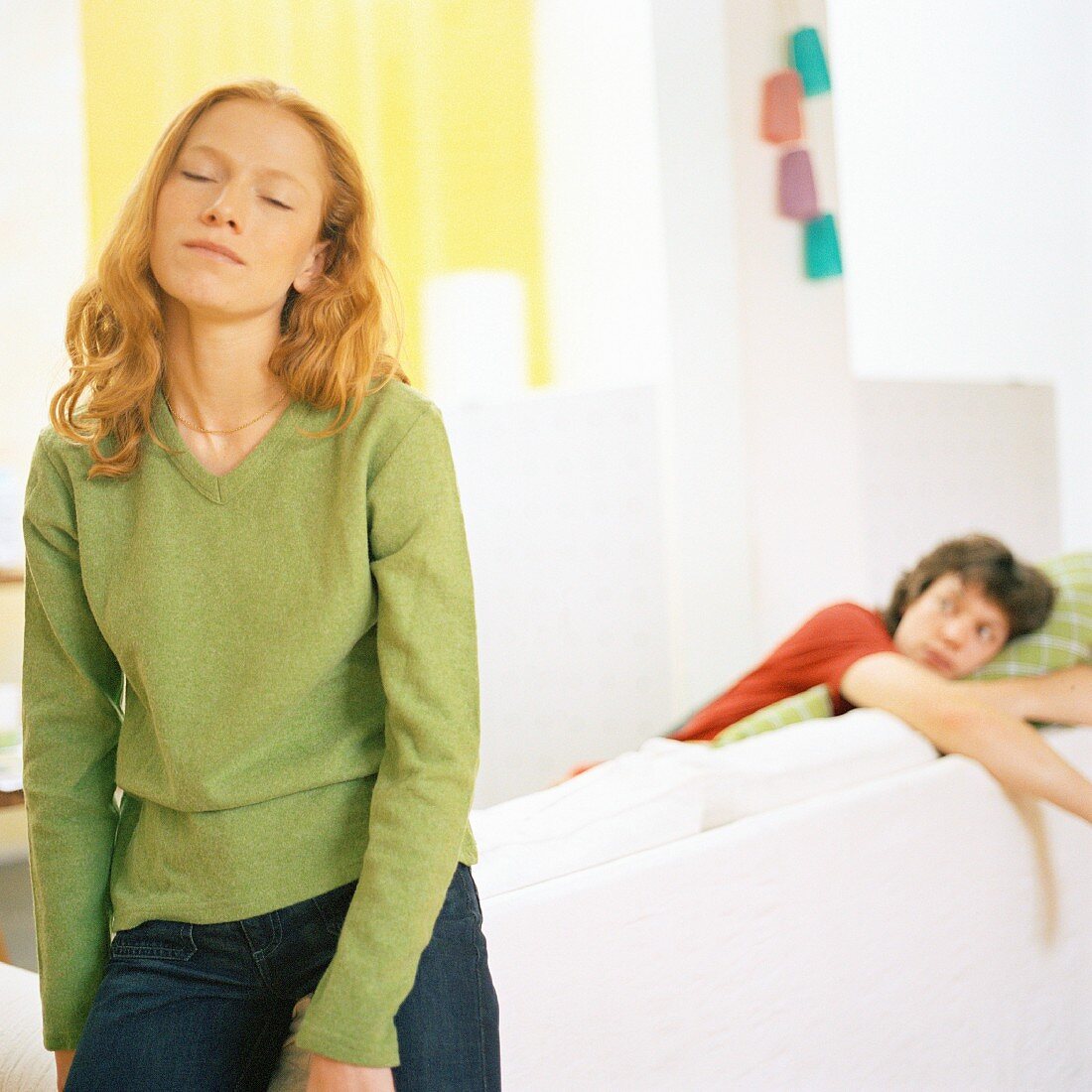 A young woman leaning on a sofa and a young man in the background lying on the sofa