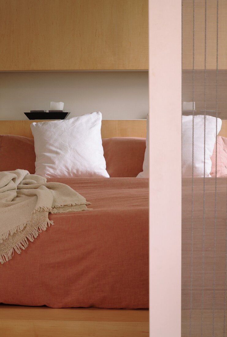 Double bed with pale bedspread against smooth wooden fronts of fitted cupboards; detail of sliding door with translucent bamboo blind