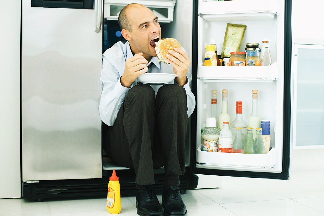Man sitting in front of open refrigerator, eating hamburger
