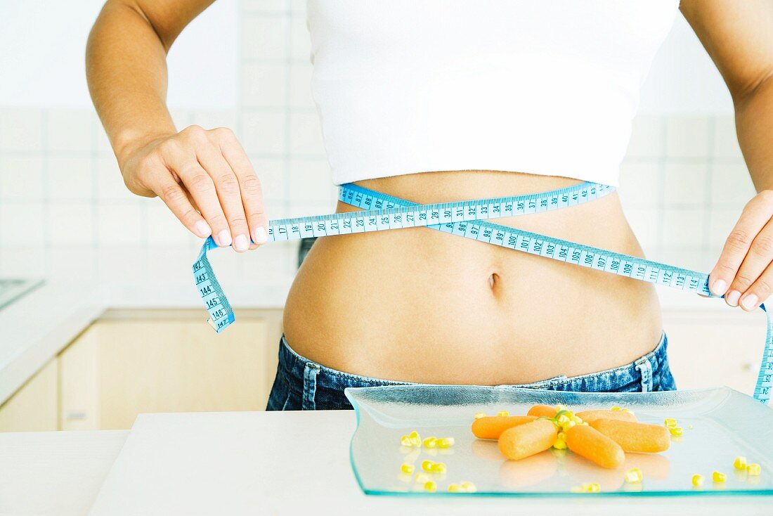 Young woman holding measuring tape around bare abdomen, plate of veggies in the foreground