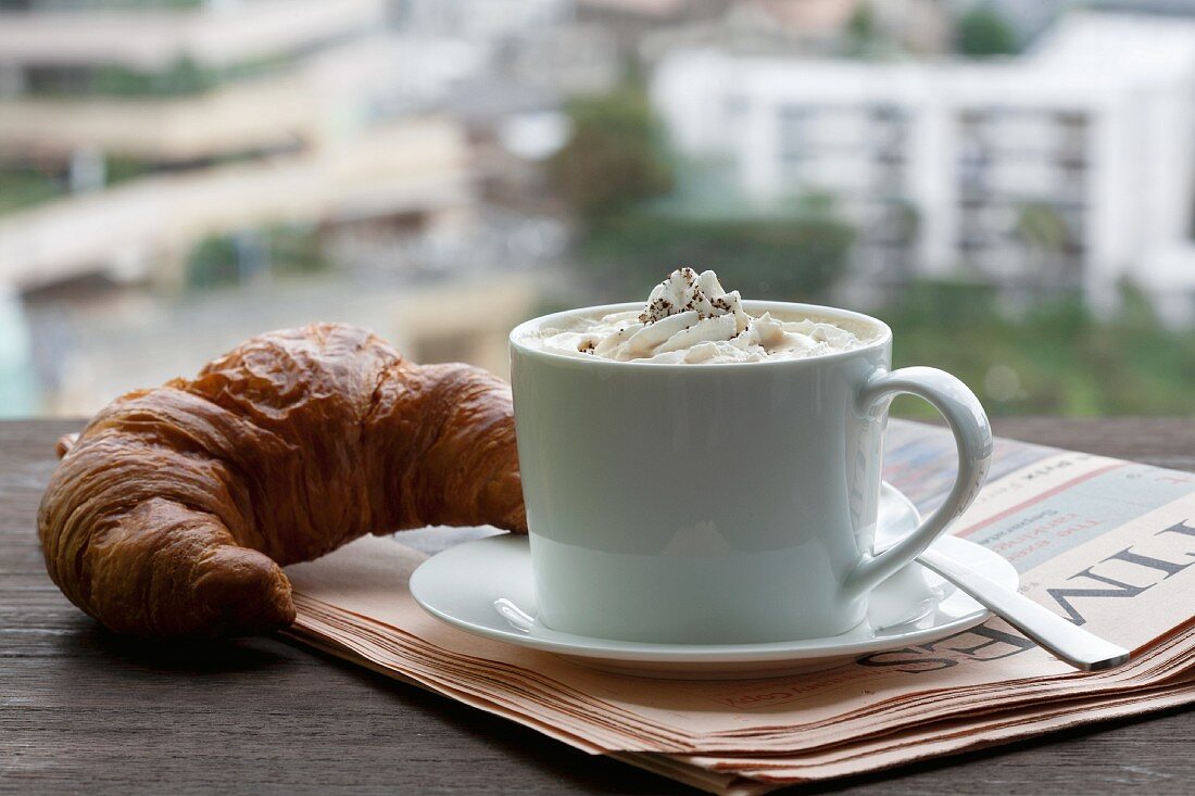 Cappuccino mit Croissant auf Zeitungspapier vor dem Fenster