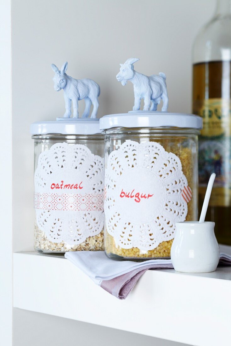 Doilies, labelled and decorated with patterned tape, stuck on storage jars with small, white china figurines on lids