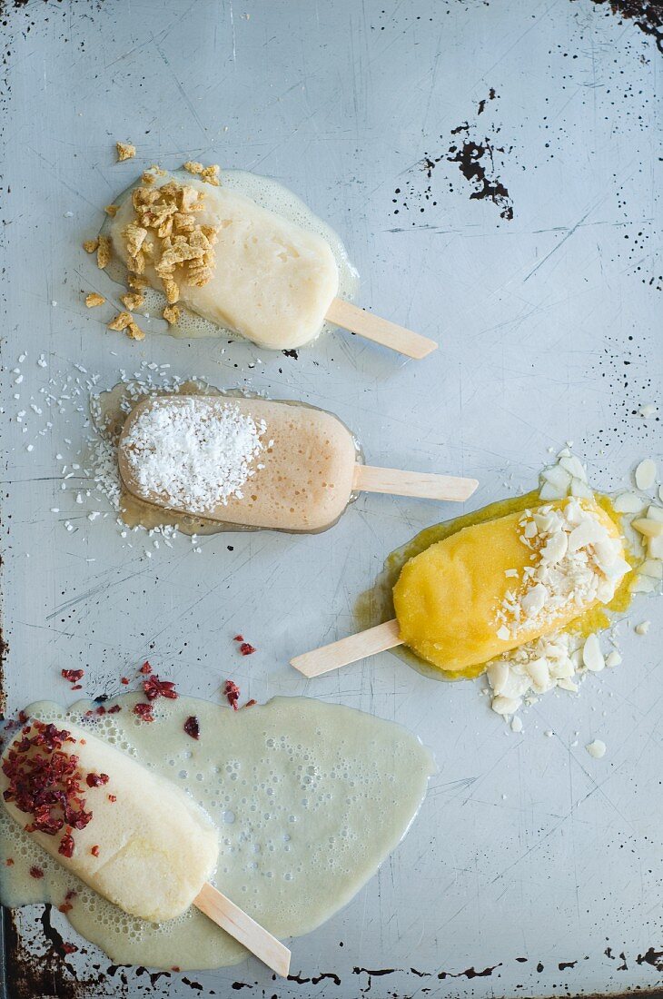 Four Ice Cream Popsicles Sprinkled with Assorted Toppings; Melting on a Metal Surface; From Above