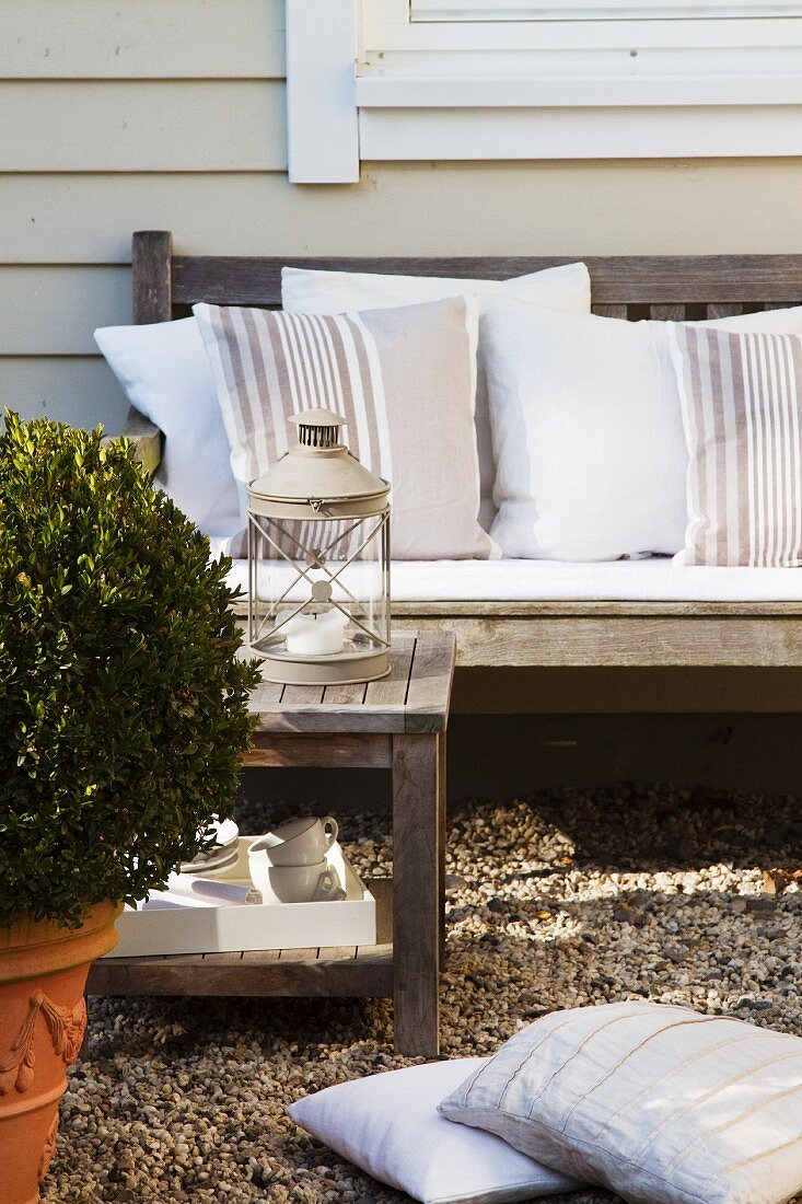 Teak bench with white cushions and candle lantern on table behind box sphere