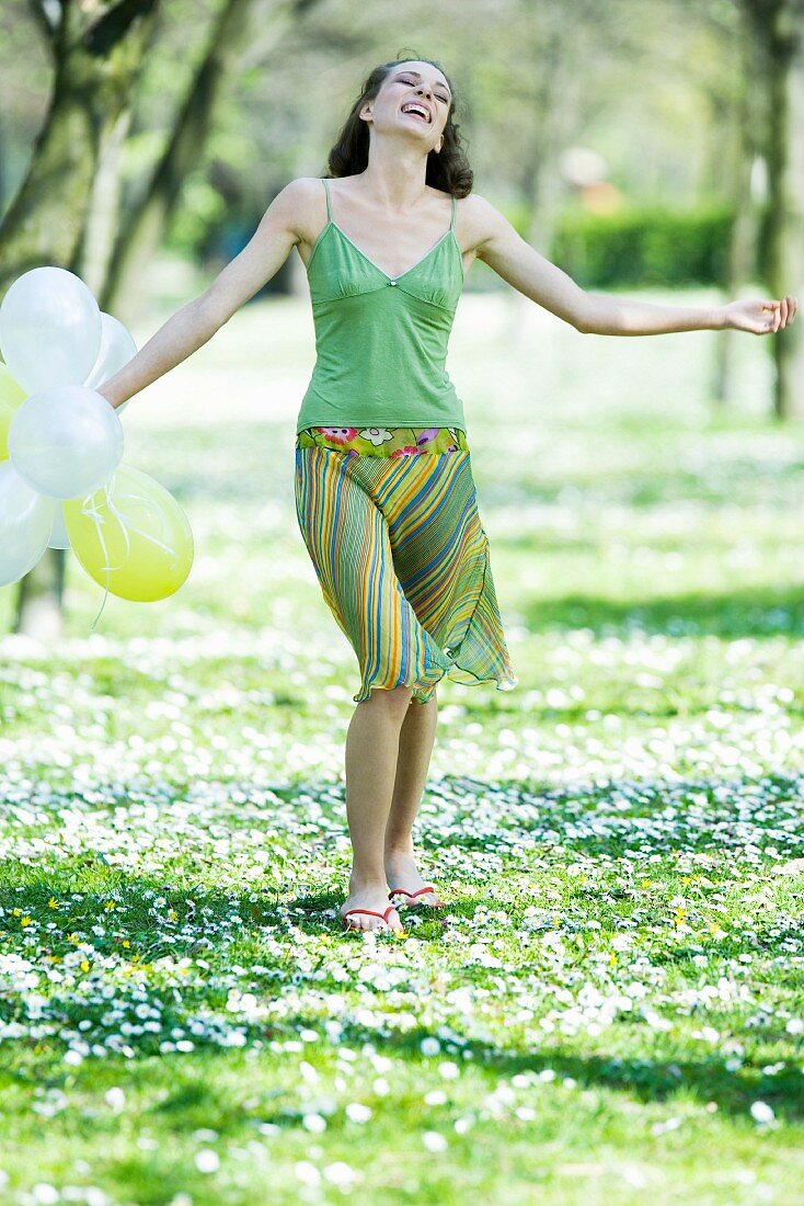 Young woman walking in meadow, holding balloons, arms out, smiling