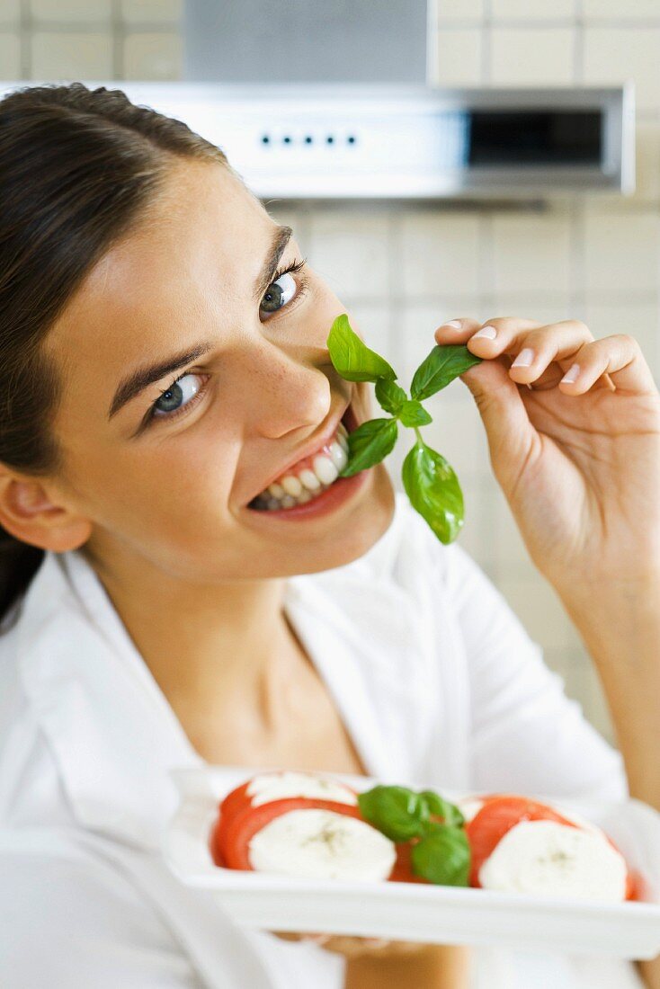 Woman biting into sprig of basil, holding plate of Caprese salad
